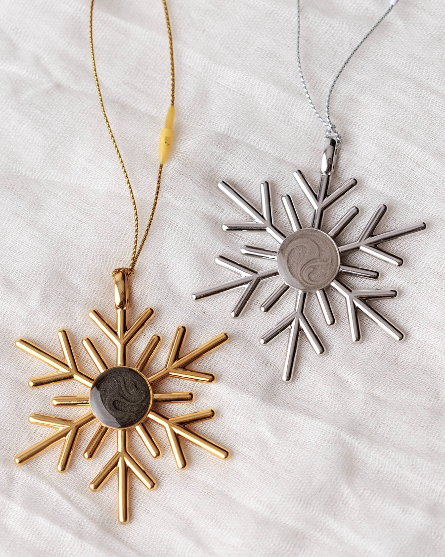 Close By Me's 2025 Snowflake Cremation Ornament in gold (left) and silver (right) on a light fabric background.