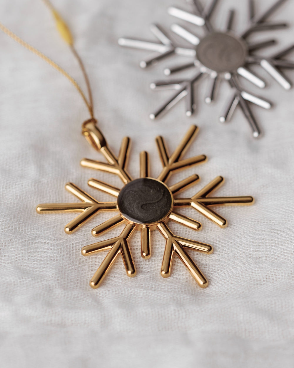 Close By Me's 2025 Snowflake Cremation Ornament in gold (centered and in focus) and in silver (back right) on a light fabric background.