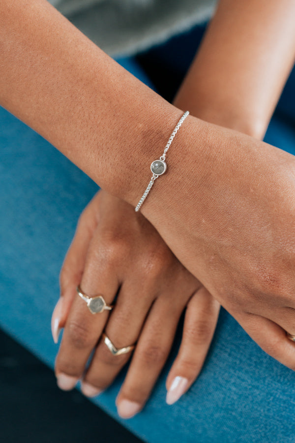 A tan model wearing a grey sweater shows the bolo chain bracelet in sterling silver on one wrist overlapping the other arm with cremation rings being worn