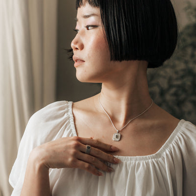 A young woman poses with her head turned to the side and hand resting gently on her chest while she wears Close By Me's Tessa Cremation Necklace and matching ring.