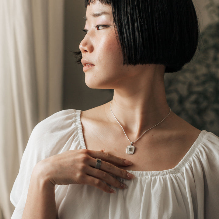 A young woman poses with her head turned to the side and hand resting gently on her chest while she wears Close By Me's Tessa Cremation Necklace and matching ring.