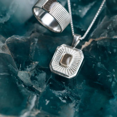 A close-up, overhead view of Close By Me's Tessa Cremation Necklace and matching ring set on a dark green agate-style backdrop.