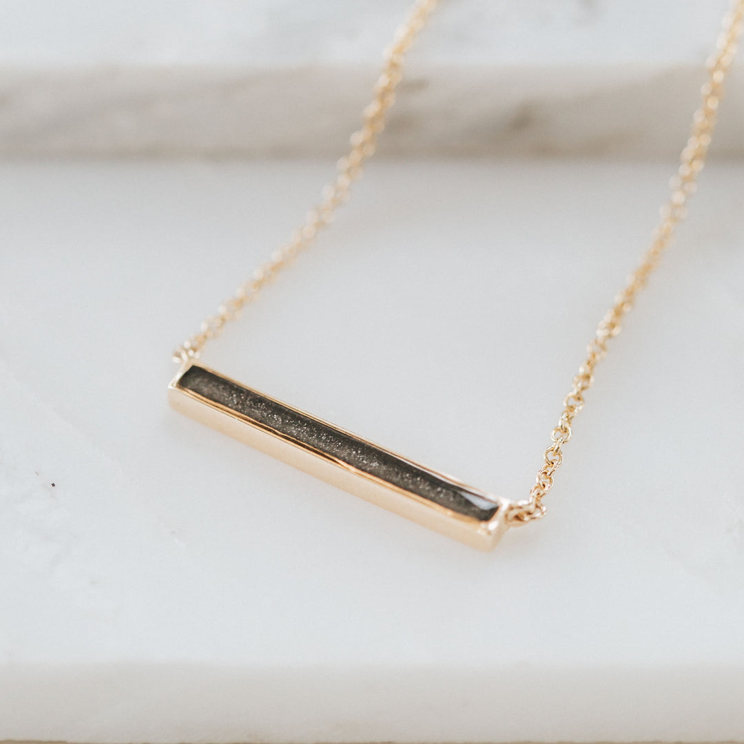 A memorial necklace with an attached chain and a thin lateral bar design in 14k yellow gold lying flat against a white background
