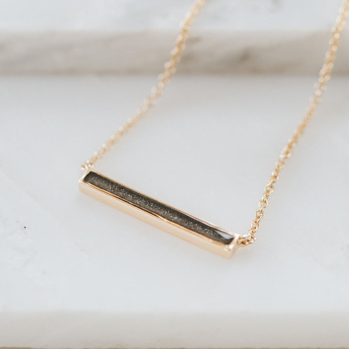 A memorial necklace with an attached chain and a thin lateral bar design in 14k yellow gold lying flat against a white background