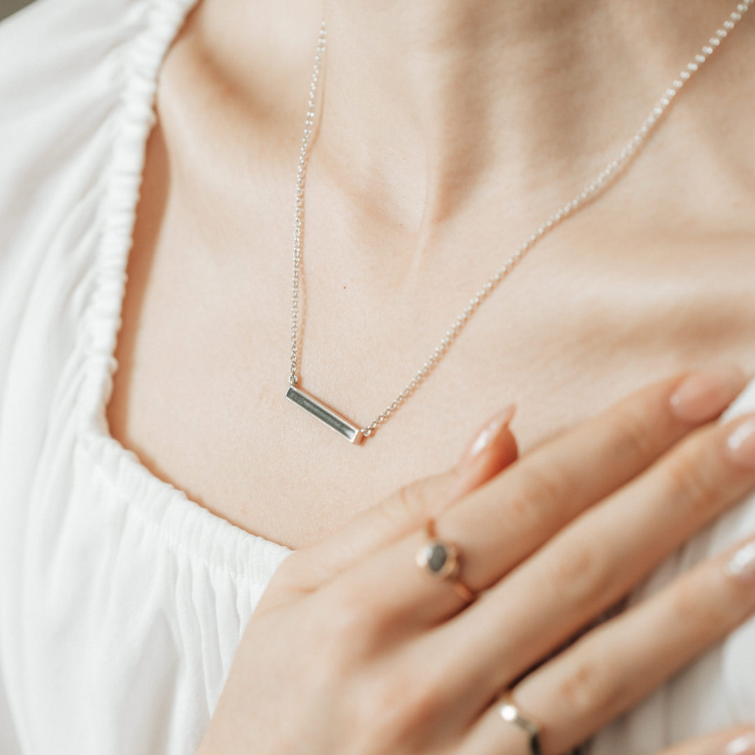 A thin lateral bar pendant memorial with an attached chain in sterling silver being worn by a model in a white top