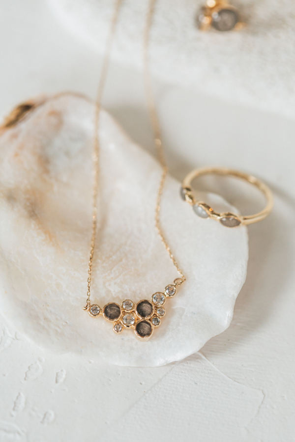 This photo shows a close up of several gold jewelry designs with ashes, set by close by me jewelry; centered in this photo is a the White Sapphire Cluster Cremation Pendant in 14K Yellow Gold resting on a white shell against a white background