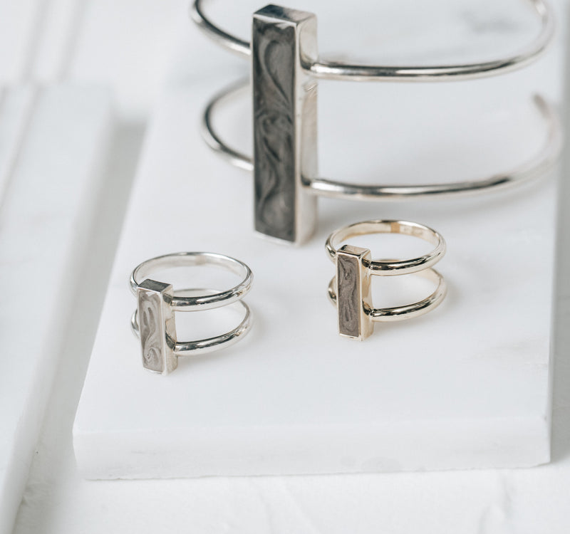 This is a stylized photo showing several pieces of Sterling Silver Cremation jewelry by close by me jewelry on a white flat stone. The Bar Cremation Bracelet and Ashes Rings are displayed upright.