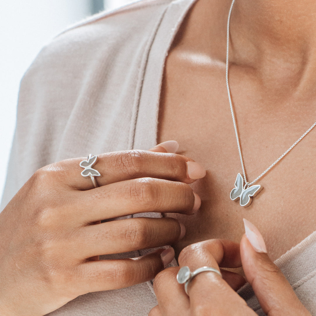Pictured here is a photo of a warm-skinned model wearing the Sterling Silver Butterfly collection by close by me jewelry. The Butterfly Ashes Ring is worn on the index finger of her left hand and held close to her chest where the Butterfly Ashes Pendant sits. Another ring is worn on the index finger of her right hand but is blurred slightly.
