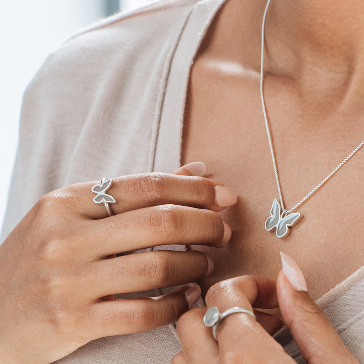 This photo shows a model with medium-warm skin wearing the Sterling Silver Butterfly Cremation collection by close by me jewelry. She wears the Butterfly Ashes Ring on her left index finger, which she holds close to the Butterfly Pendant that rests against her chest.
