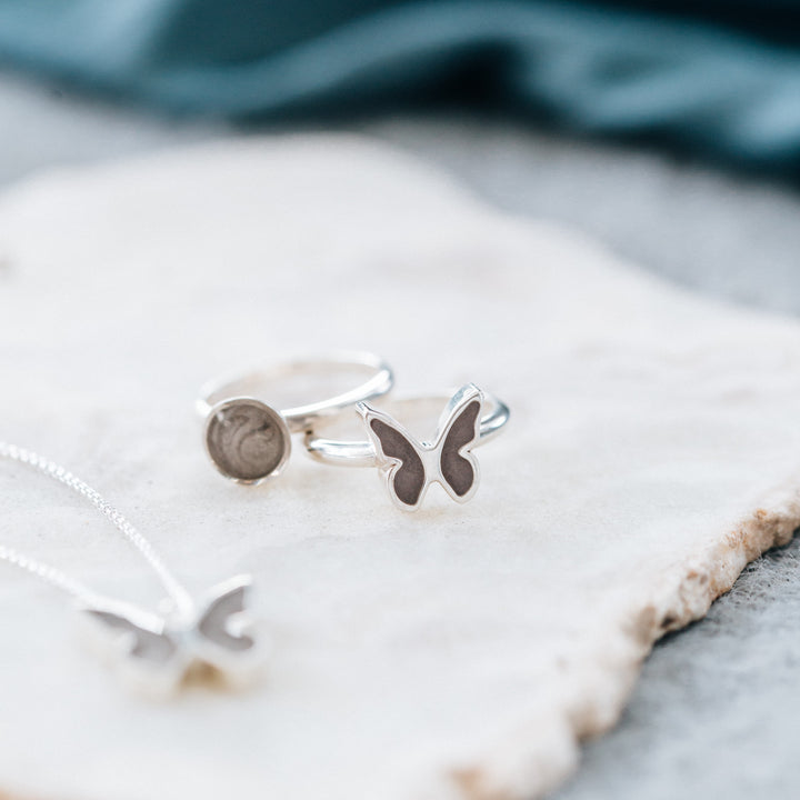 This photo shows several pieces of Sterling SIilver Ashes jewelry by close by me jewelry. The foreground and background are blurred but show a Butterfly Pendant and white stone upon which the jewelry rests. The Butterfly and Signature Lotus Stackable Band Ashes Rings face forward and rest against each other.