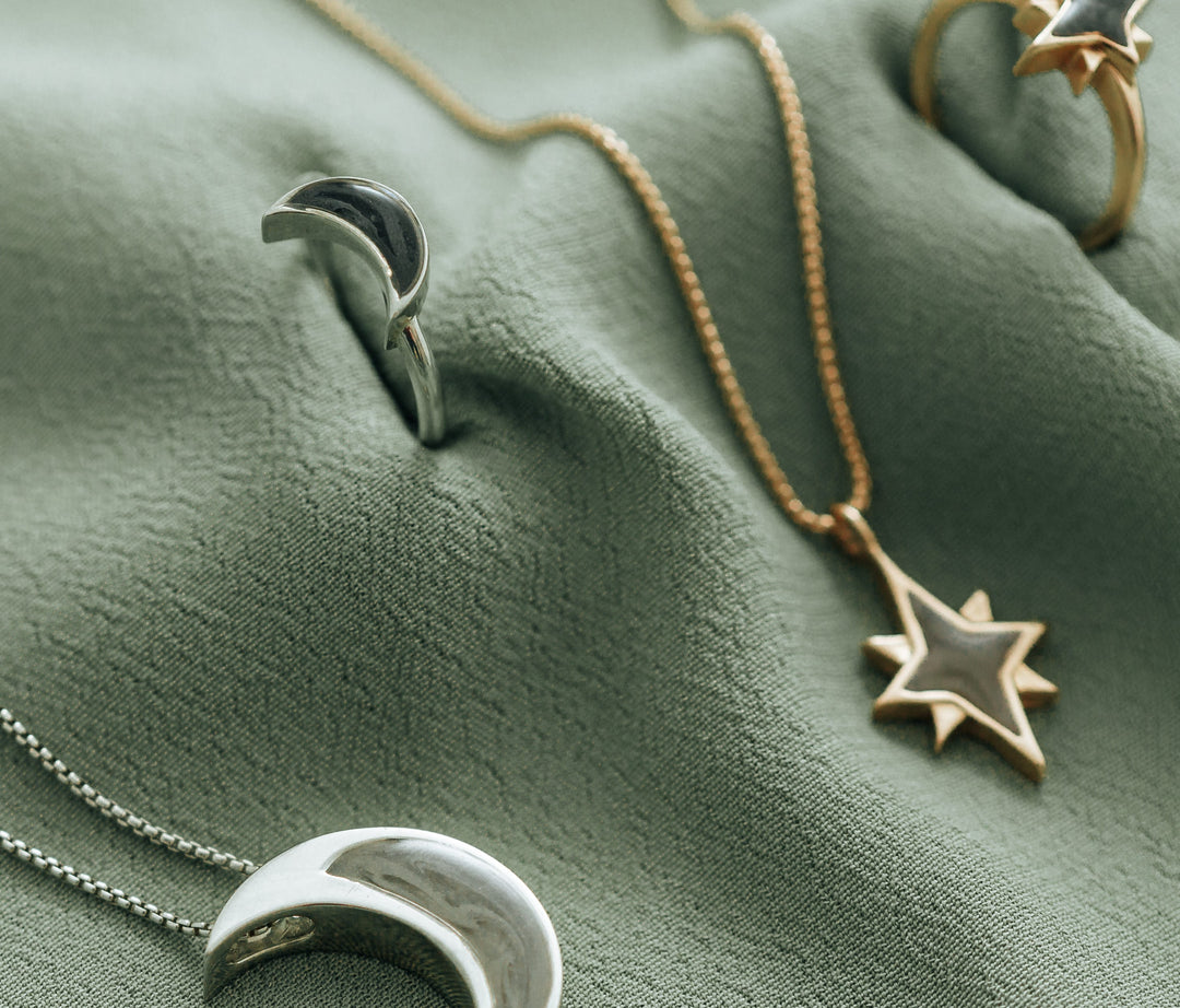 This photo shows several pieces of mixed metal cremation jewelry lying on a safe green textured cloth. The pieces are shown in both Sterling Silver and 14K Yellow Gold, but the star pieces are blurry. The Sterling Silver Crescent Moon Ring is most prominent and shows the dark gray ashes setting.