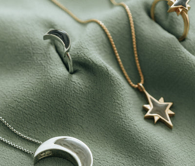 This photo shows several pieces of ashes jewelry by close by me jewelry on a textured green cloth. There are 14K Yellow Gold North Star designs that are blurry and the Sterling Silver Crescent Moon Ring is most prominent, facing up toward the camera to show its dark gray ashes setting