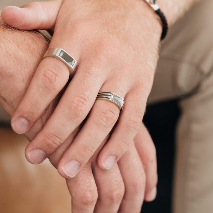 side angle of male hand wearing close by me's classic band cremation ring on index finger and ridged band rectangle signet cremation ring on ring finger