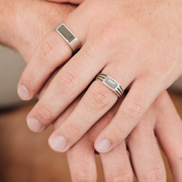 looking down at male hand wearing close by me's classic band cremation ring on index finger and ridged band rectangle signet cremation ring on ring finger