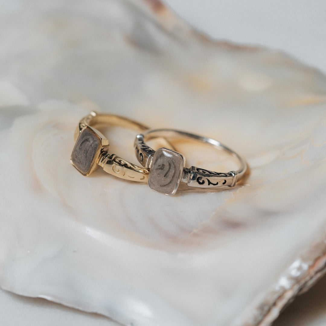 This photo shows close by me jewelry's Emerald Setting Cremation Ring design in both 14K Yellow Gold and Sterling Silver lying flat on a shell