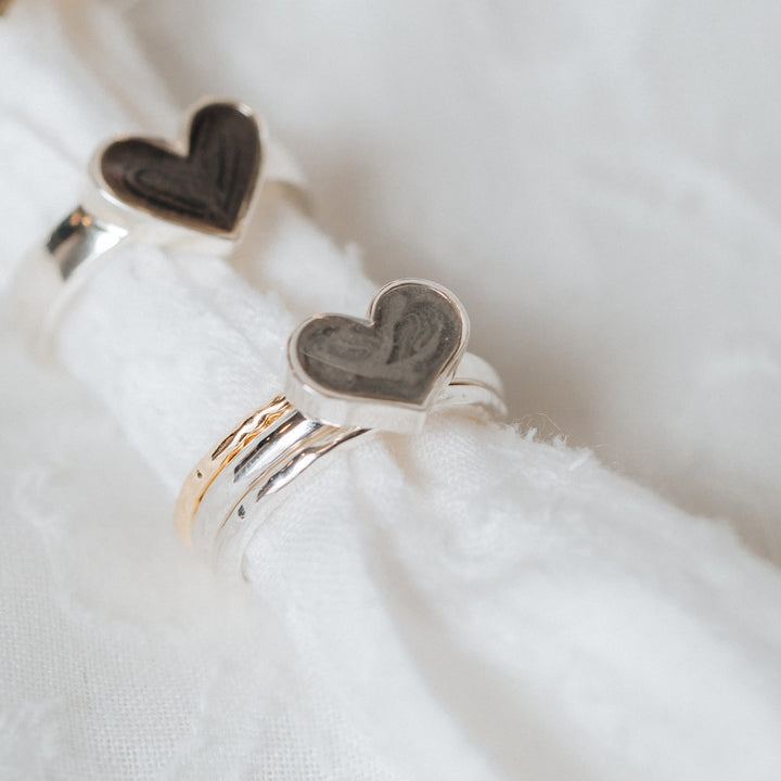 This stylized photo shows close by me jewelry's Sterling Silver Imprint Heart and Large Heart Stacking Rings in Sterling Silver with a piece of soft white cloth threaded through the bands. The Large Heart Stacking Ashes Ring is paired with Textured Companion Rings in both Sterling Silver and 14K Yellow Gold to create a stacking ashes ring set.
