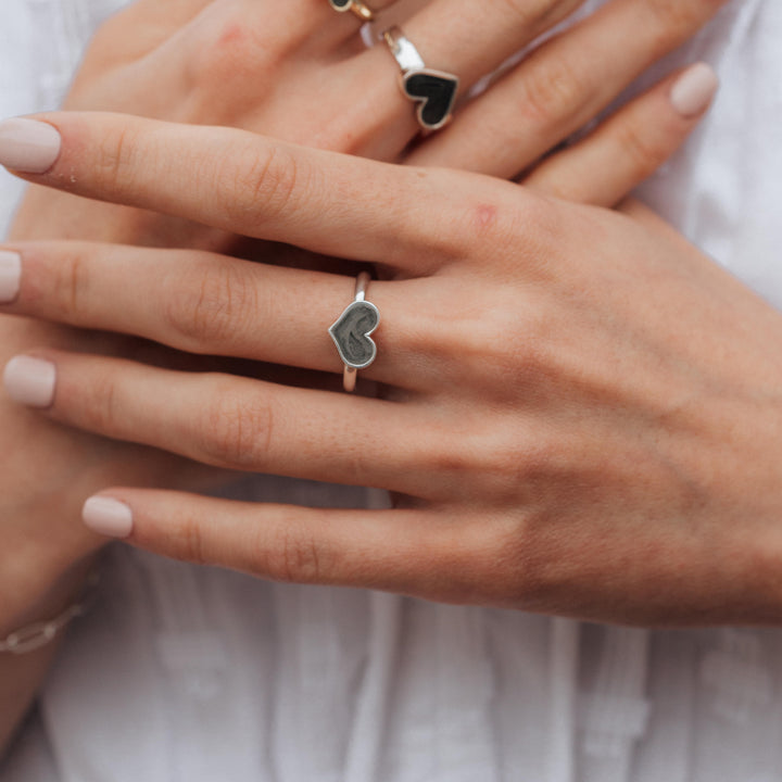 This photo shows a model wearing several Heart Shaped Cremation Ring designs on her fingers by close by me jewelry. The hand in front shows the Large Heart Stacking Cremation Ring in Sterling Silver and her right hand, which she holds behind, is wearing the Imprint Heart Ashes Ring in Sterling Silver.