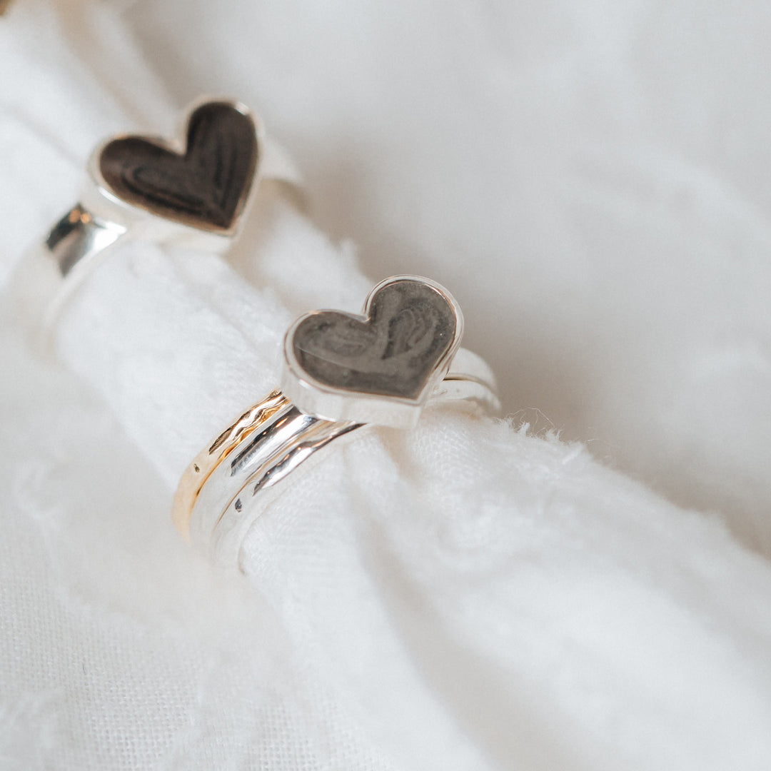 This is a stylized photo of the Sterling Silver Large Heart Stacking and Imprint Heart Cremation Rings in Sterling Silver on a piece of soft white cloth. The Large Heart Stacking Cremation Ring is paired with two Companion Stacking Rings, one in 14K Yellow Gold with a Textured finish and one with a smooth finish.