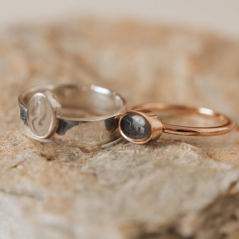 Pictured here is a 14K Rose Gold Lateral Oval Cremation Stacking Ring designed by close by me jewelry lying flat next to a Sterling Silver ring with ashes