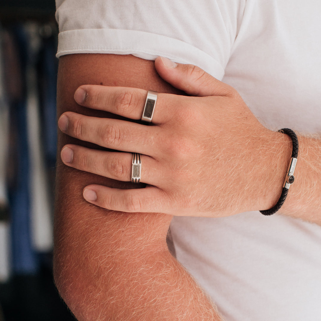 Pictured here is a male model wearing close by me jewelry's Men's Classic Band and Ridged Rectangle Signet Cremation Rings in Sterling Silver and the Leather Cord Cremation Bracelet