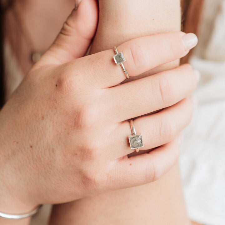 This photo shows a model wearing several rings with ashes by close by me jewelry in Sterling Silver. On her index finger, she wears the Small Square Stacking Cremains Ring design..