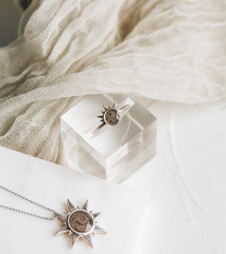 This stylized photo shows the Sunrise Ashes Ring and Sun Ashes Pendant in Sterling Silver designed by close by me jewelry positioned on a multi-textured white background
