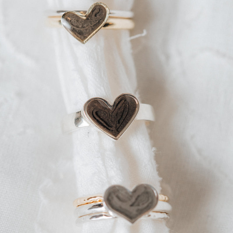 This photo shows three different Heart Cremation Ring designs by close by me jewelry on a piece of soft white cloth. The Imprint Heart Ring in Sterling Silver is in the center.