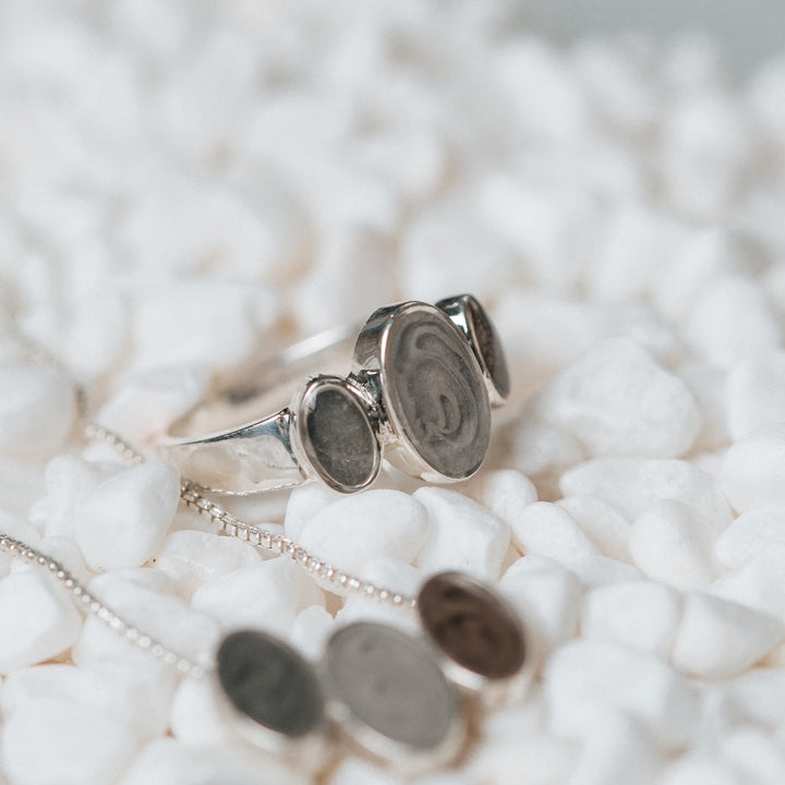 Pictured here is the Triple Oval Cremains Ring in Sterling Silver lying amongst small white pebbles